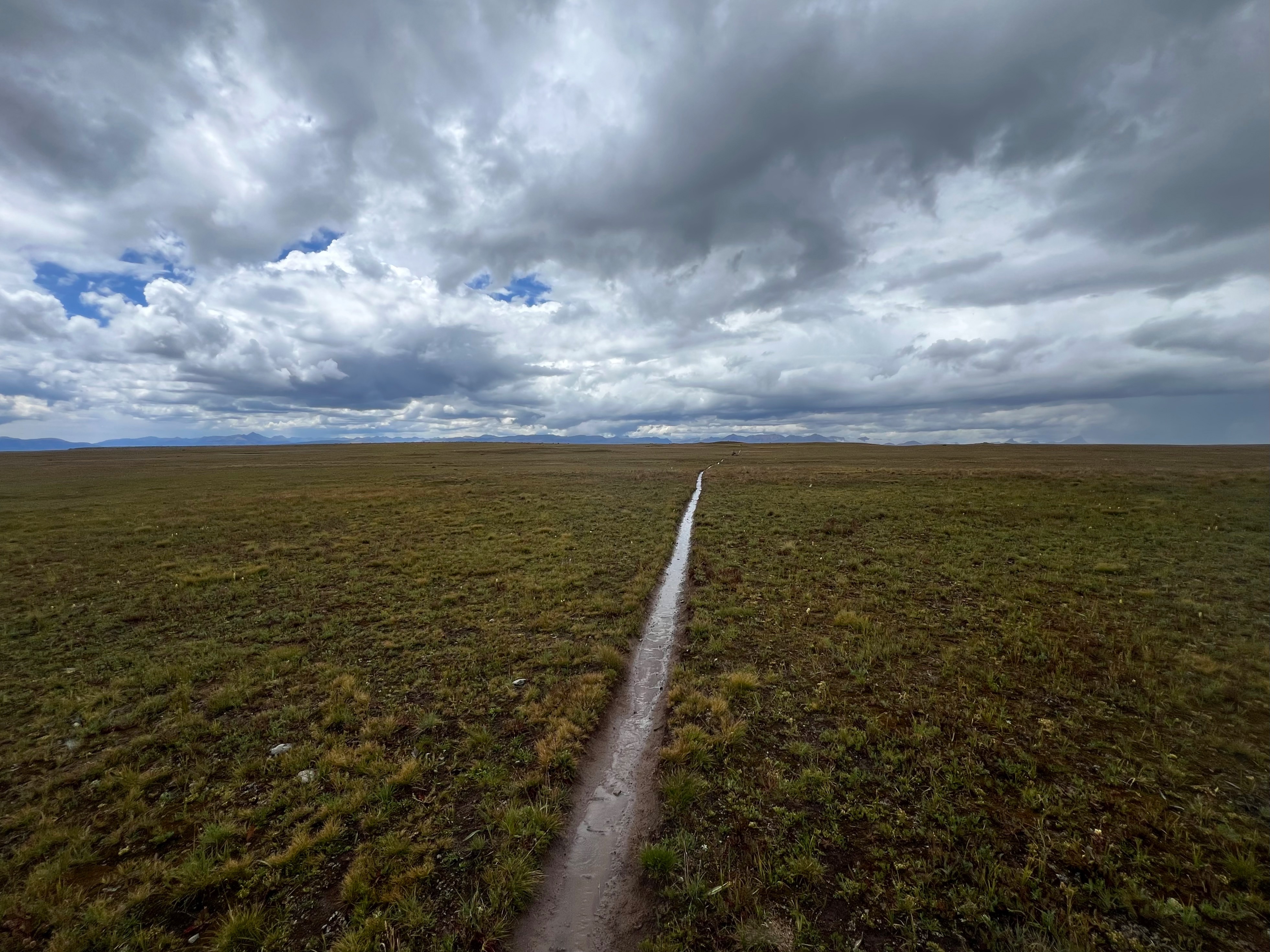 A landscape photo of a grassy plain with a long, muddy line through it and a white and gray cloudy sky with some blue peeking out.